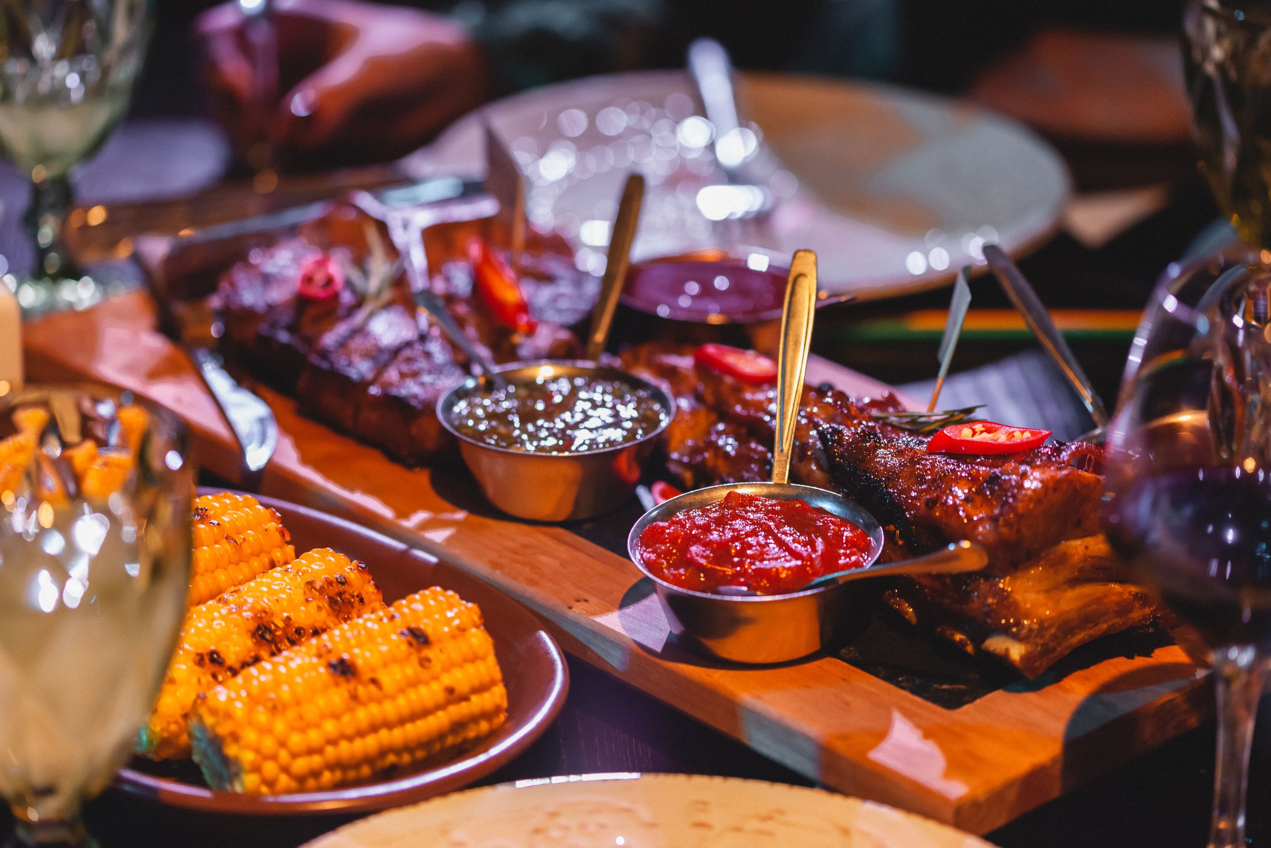Close-up of grilled ribs and corn with sauces on a wooden board, perfect for barbecue lovers.