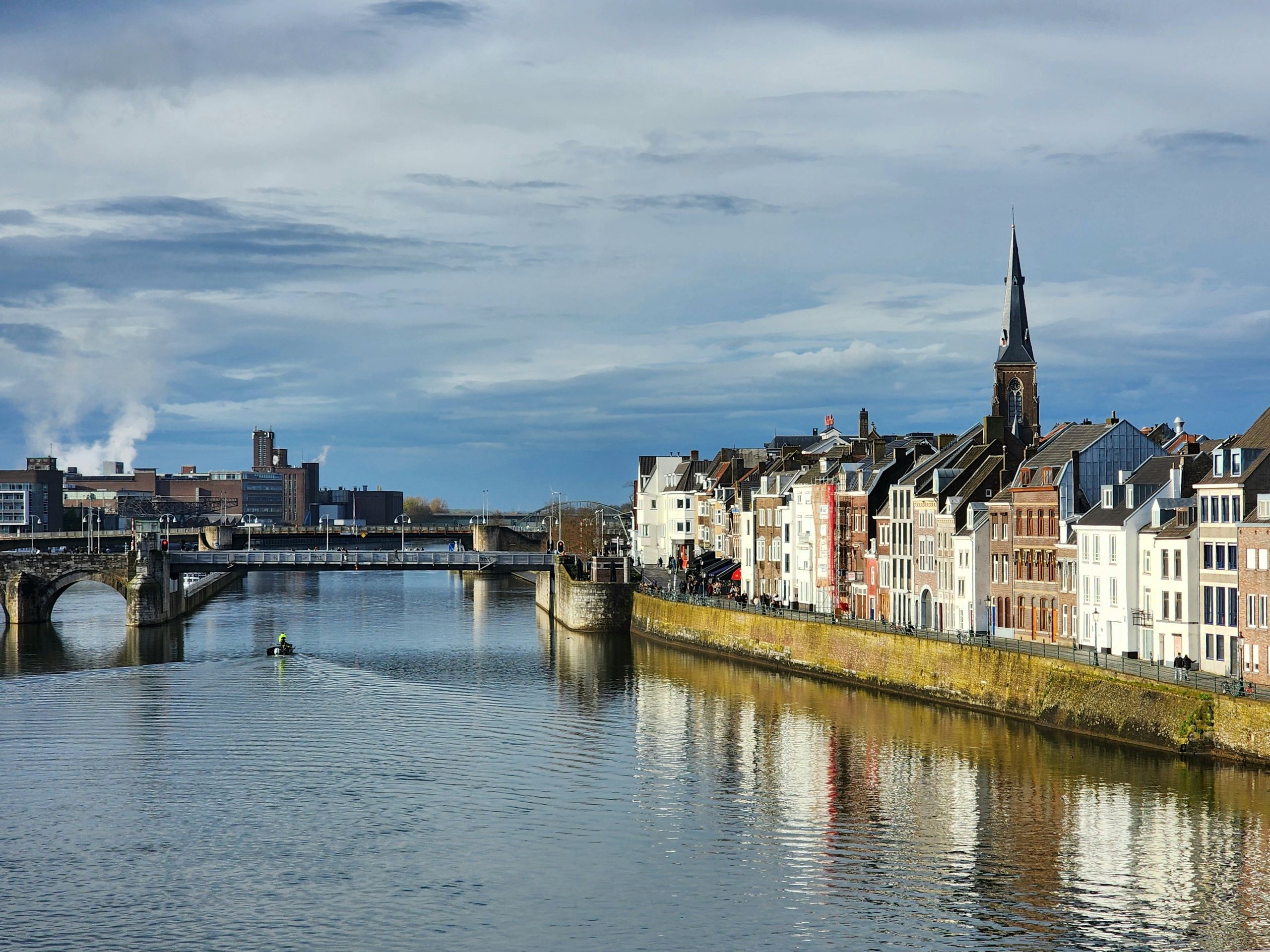 View of Maastricht's historic townhouses and bridge along the Meuse River under a cloudy sky.