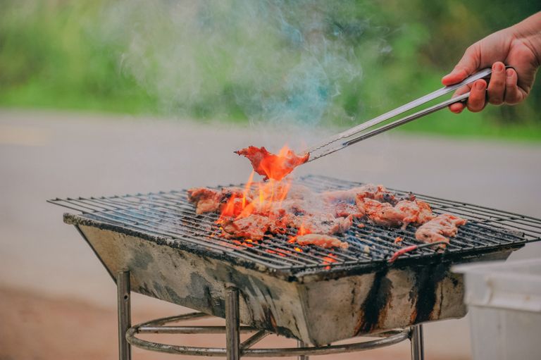 Close-up of a barbecue grill outdoors with meat cooking over flames, emitting smoke.