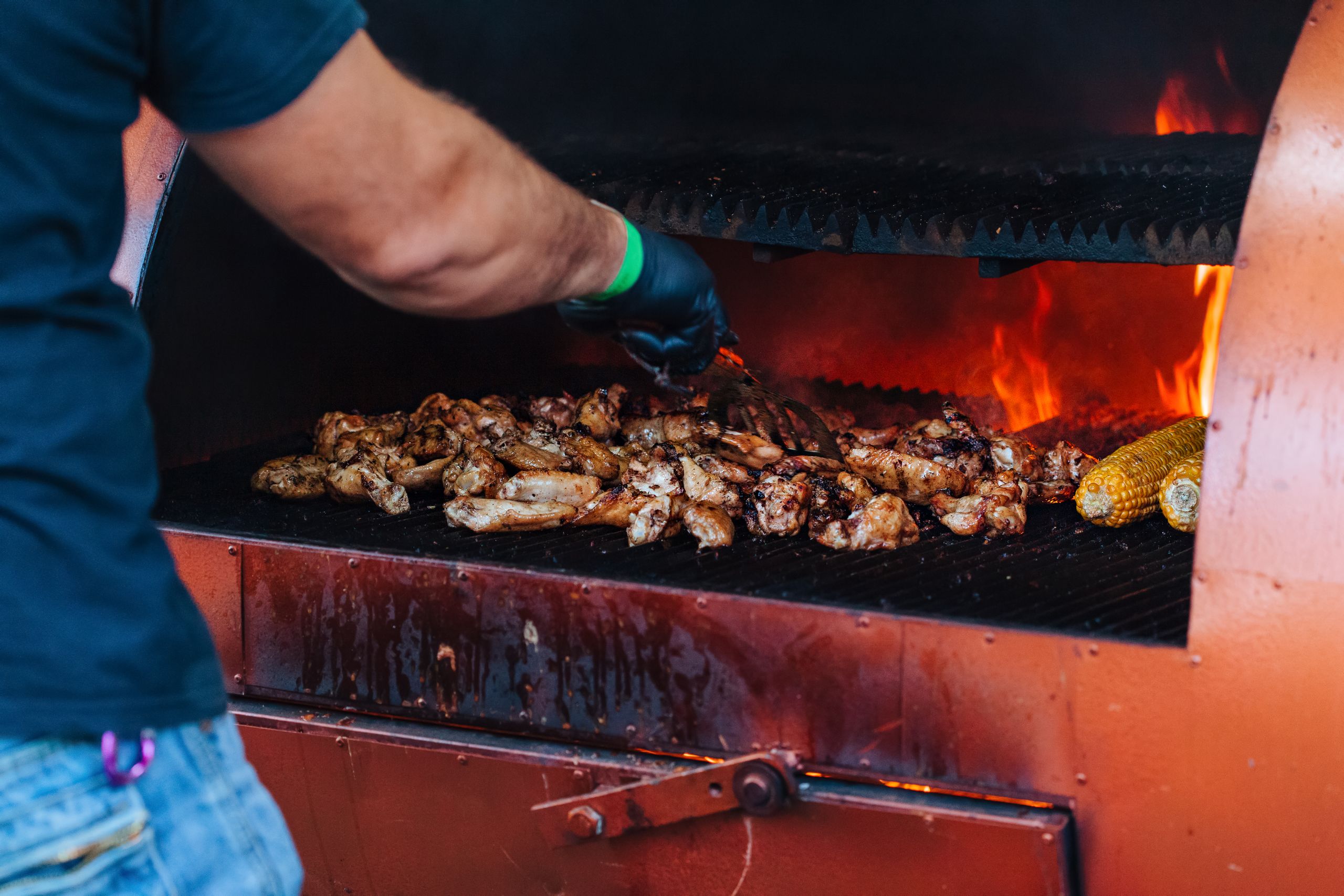 Close-up of grilling chicken wings and corn on an outdoor barbecue with flames.
